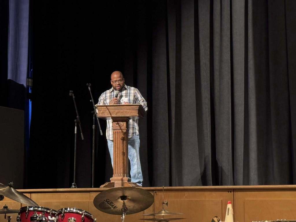 A man is standing at a podium on a stage, speaking into a microphone. A drum set can be seen nearby.
