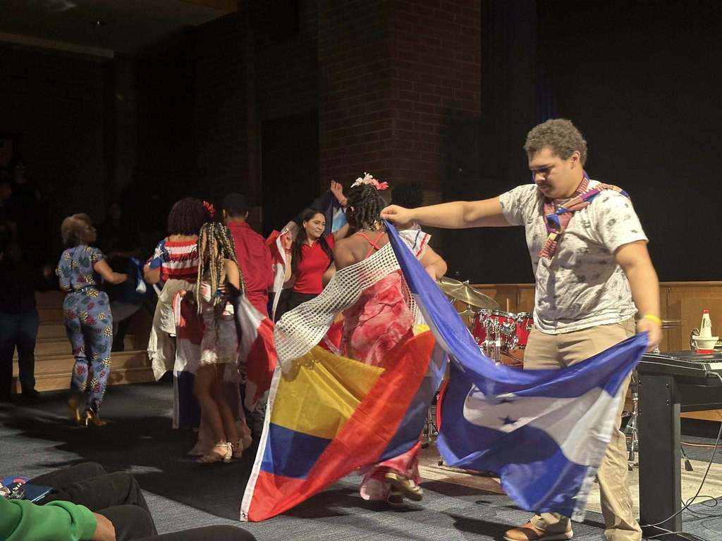 A lively dance scene in a school auditorium, featuring diverse student and staff performers in vibrant attire holding Colombian and Honduran flags.
