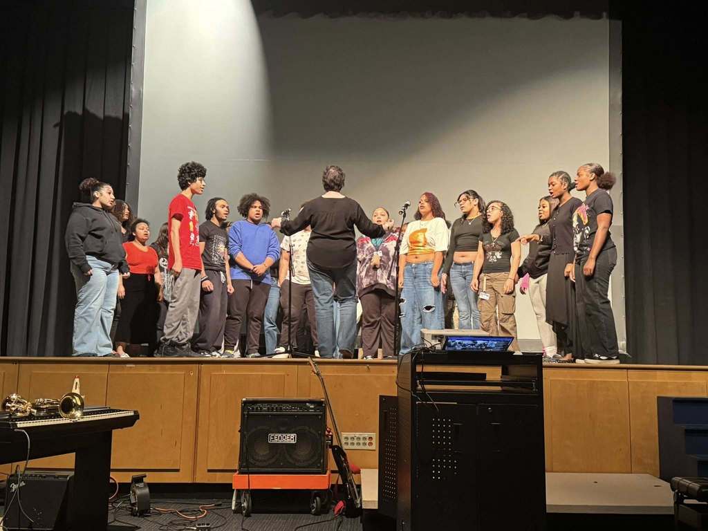 A diverse group of students are gathered on a stage singing. An instructor is standing at the center, guiding them. 