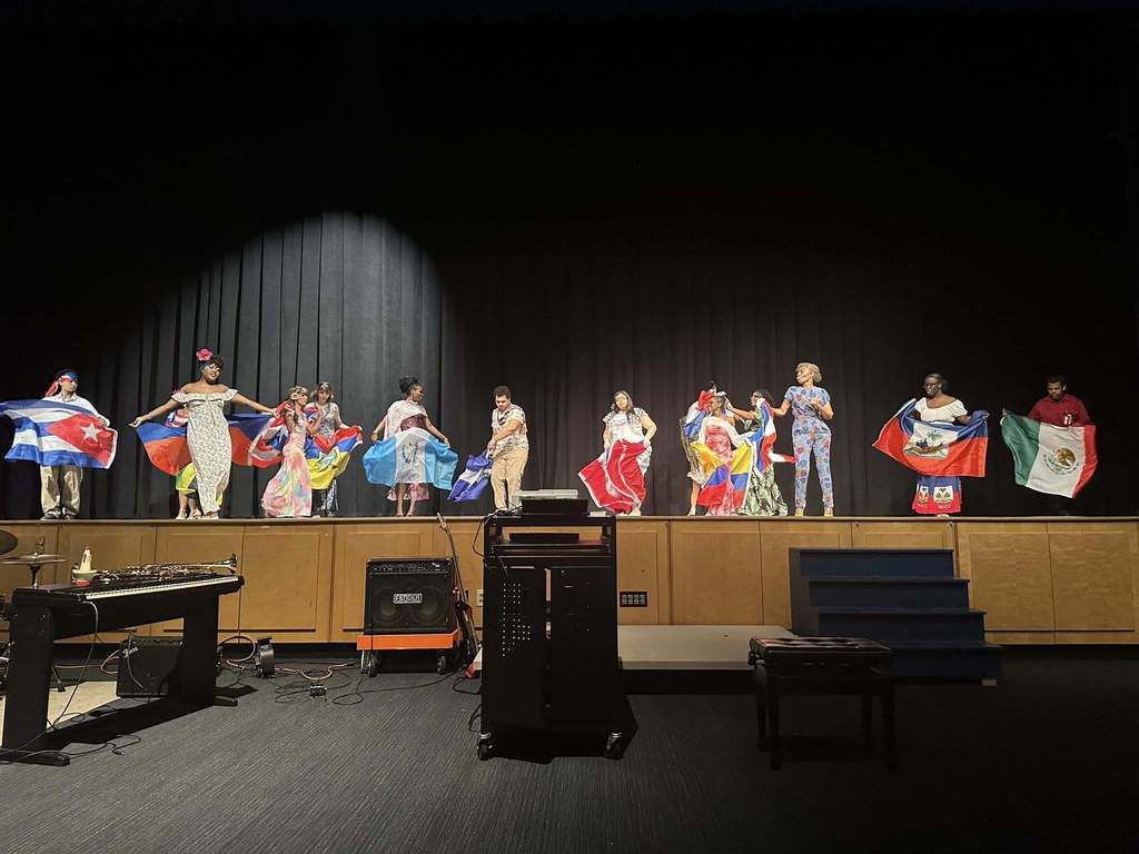 A diverse group of students and a staff member are standing on stage holding various national flags. 