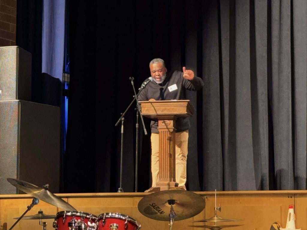 A man is standing at a podium on a stage, speaking passionately into a microphone. A drum set can be seen nearby.