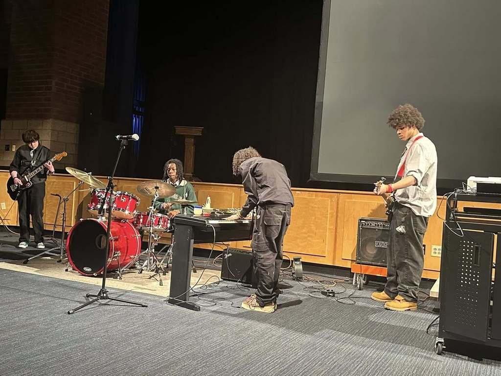 Four student musicians are performing on stage; one is playing the bass guitar, one is playing on drums, another student is adjusting a keyboard, and one student is playing electric guitar. 