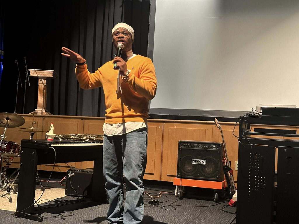 A man is speaking into a microphone near a stage. Musical instruments and equipment can be seen nearby.