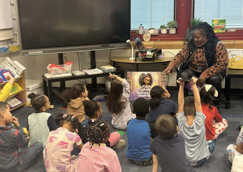 Local community member and author, Dr. Sherry Washington, is pointing to a student in a classroom of students who are sitting on a carpet. A few of the students are raising their hands in the air.