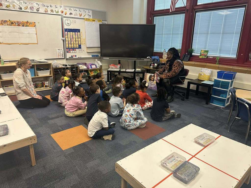 Local community member and author, Dr. Sherry Washington, is reading to a classroom of students who are sitting on a carpet. A district staff member can be seen sitting behind the students on the carpet.