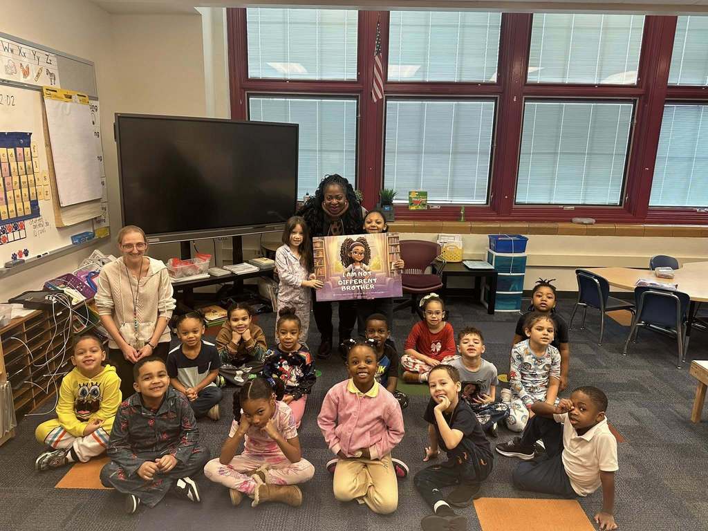 Local community member and author, Dr. Sherry Washington, a district staff member and a classroom of students are sitting and standing in a school classroom. Two of the students are holding a poster that says "I am not different brother." 