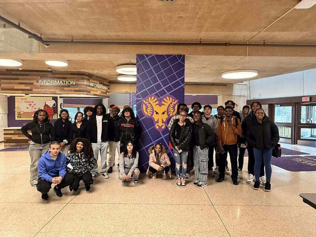 A large group of students are standing in a college lobby with a purple and yellow ram emblem behind them. 