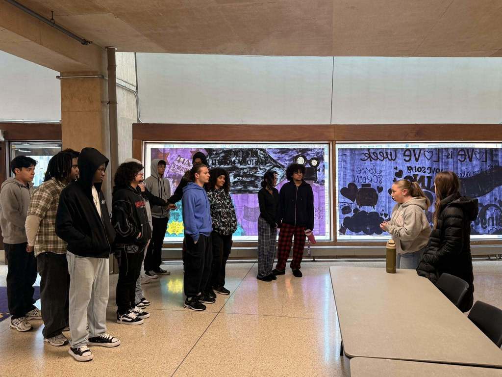 A group of students are standing in a college lobby facing two tour guides who are speaking to the group. Colorful posters with text and art are displayed on the walls in the background.