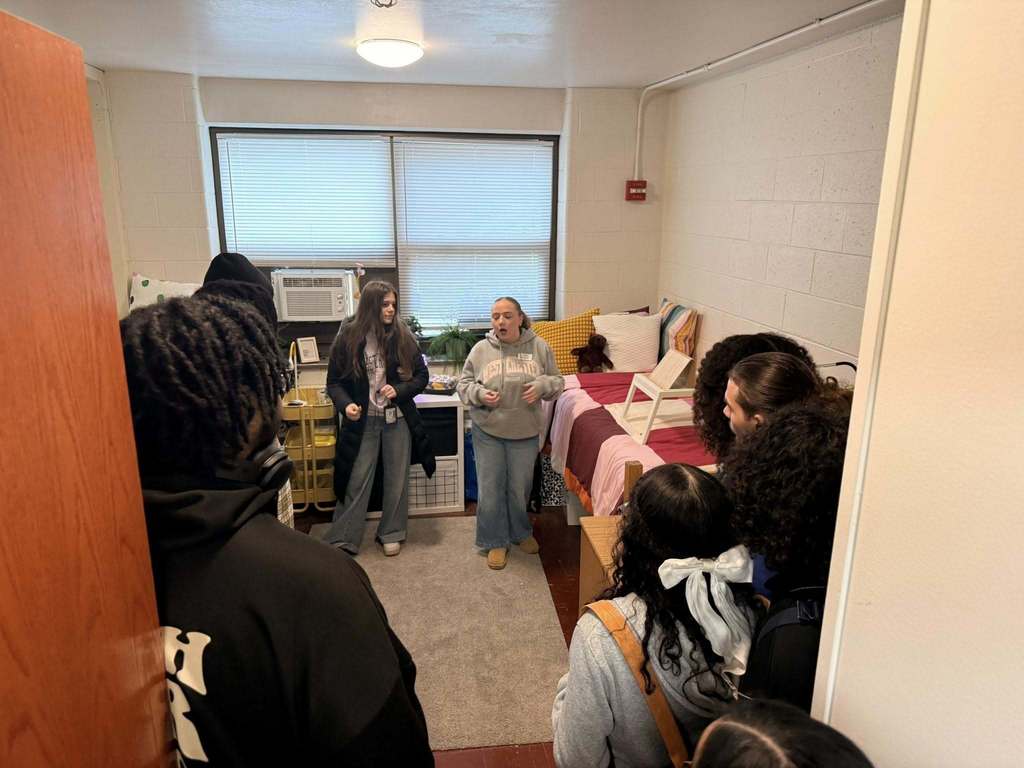 A group of students are standing in a dorm room, looking at two tour guides who are at the front of the room, speaking to the group. The room has a bed with colorful blankets and a window with blinds.
