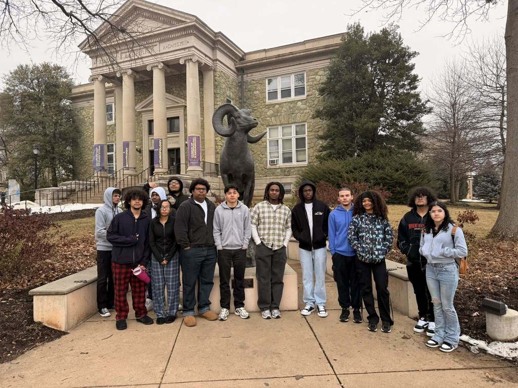 A large group of students are standing outdoors in front of a historic building with large columns, featuring a statue of a ram. 