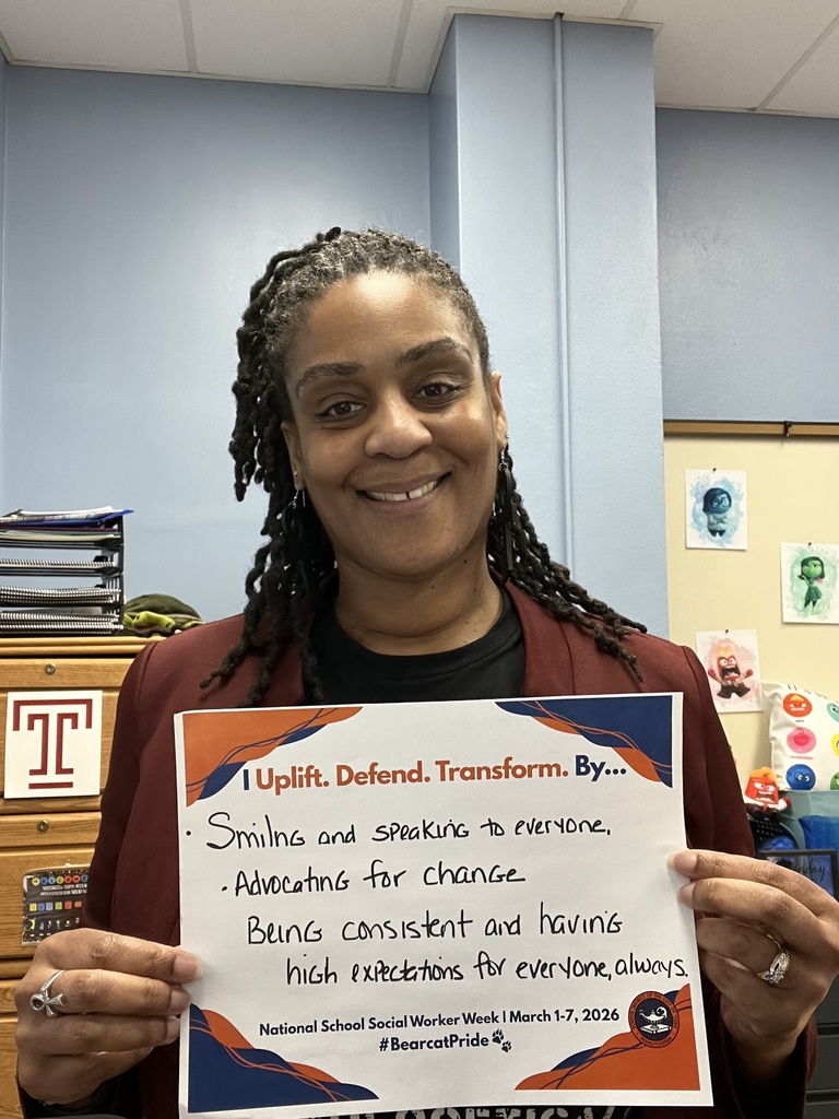 A district staff member is holding a sign that says "I Uplift. Defend. Transform. By... Smiling and speaking to everyone, Advocating for change, Being consistent and having high expectations for everyone, always."