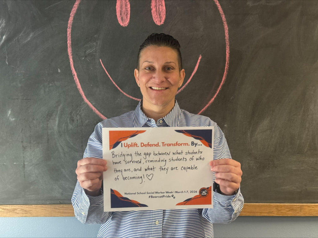 A district staff member is holding a sign that says "I Uplift. Defend. Transform. By... Bridging the gap between what students have survived, reminding students of who they are, and what they are capable of becoming!" with a heart after the exclamation point.