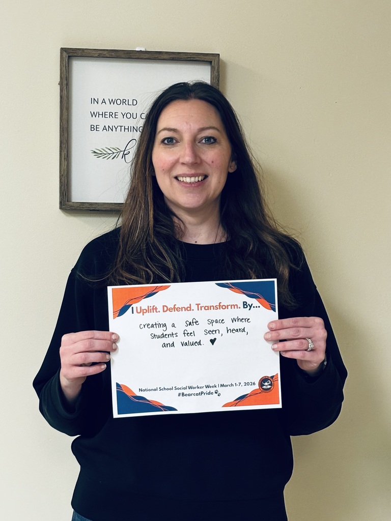 A district staff member is holding a sign that says "I Uplift. Defend. Transform. By... creating a safe space where students feel seen, heard, and valued" with a heart after the period.