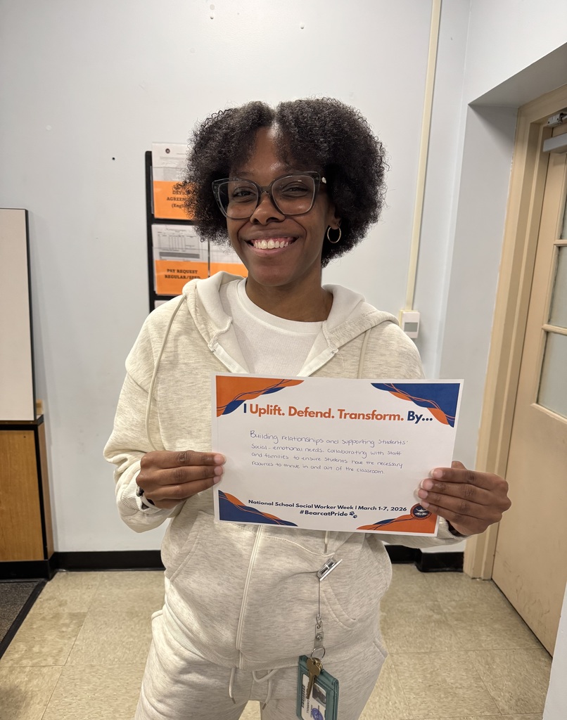 A district staff member is holding a sign that says "I Uplift. Defend. Transform. By... Building relationships and supporting students' social-emotional needs, collaborating with staff and families to ensure students have the necessary resources to thrive in and out of the classroom."