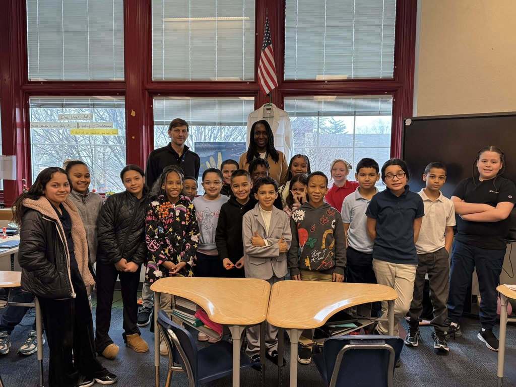 York City Mayor Sandie Walker, a district staff member and a group of students are standing behind two desks in a school classroom. An American flag can be seen hanging above them. 