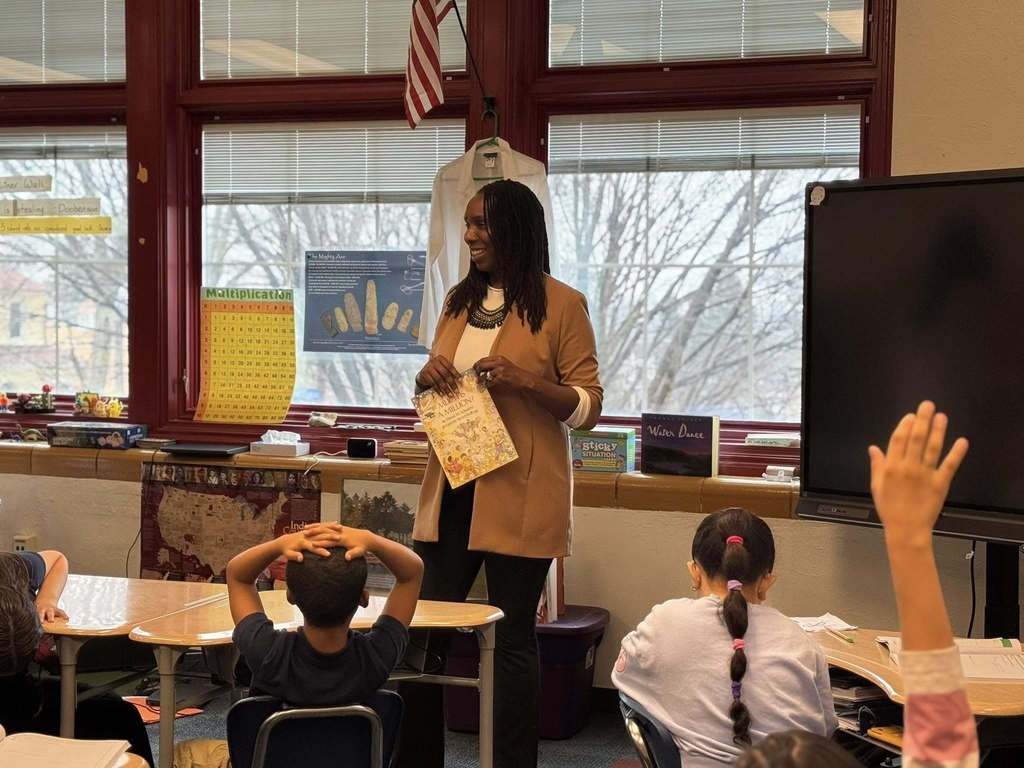 York City Mayor Sandie Walker is standing in front of a group of students in a school classroom, holding a book in her hand. The students are seated at desks, with one of the students raising their hand in the air. 