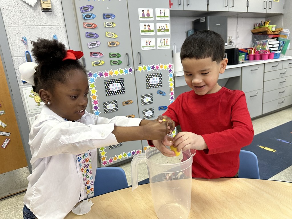 Two students are sitting at a table in a school classroom, squeezing lemons into a clear pitcher. Educational posters and storage cabinets can be seen in the background.