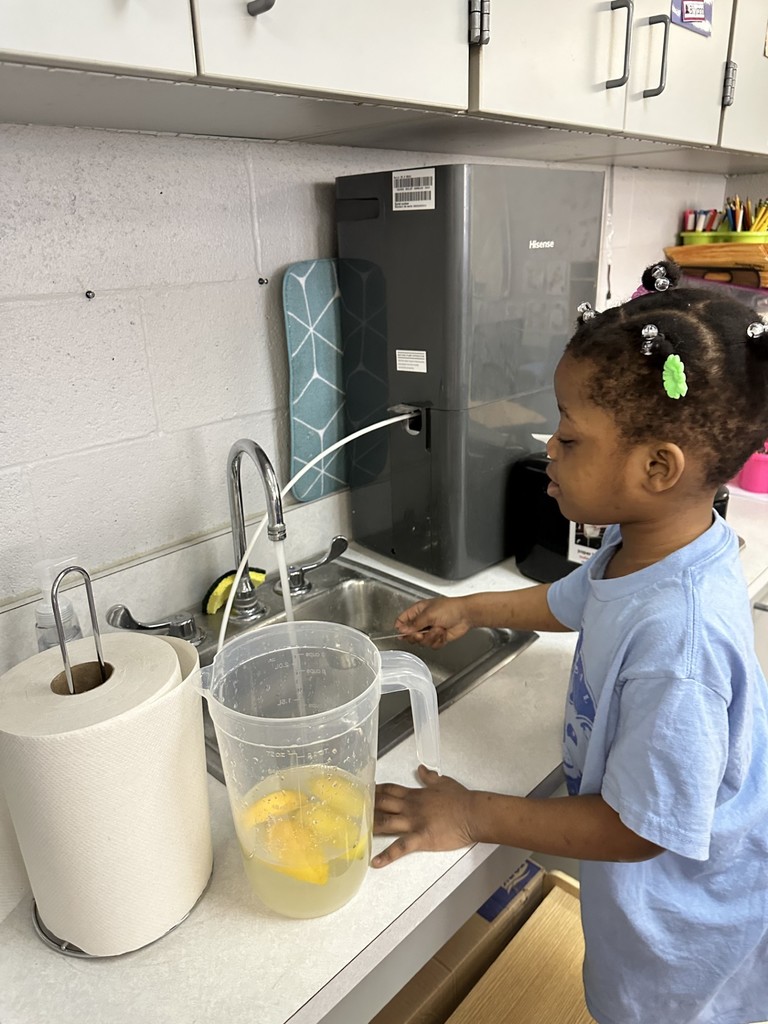 A student is filling water from a faucet into a clear pitcher with lemon slices that is sitting on a counter. A paper towel roll is nearby. 