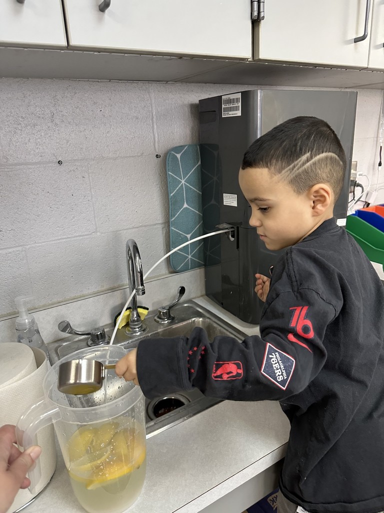 A student is filling water from a faucet into a clear pitcher with lemon slices that is sitting on a counter. A paper towel roll is nearby. 