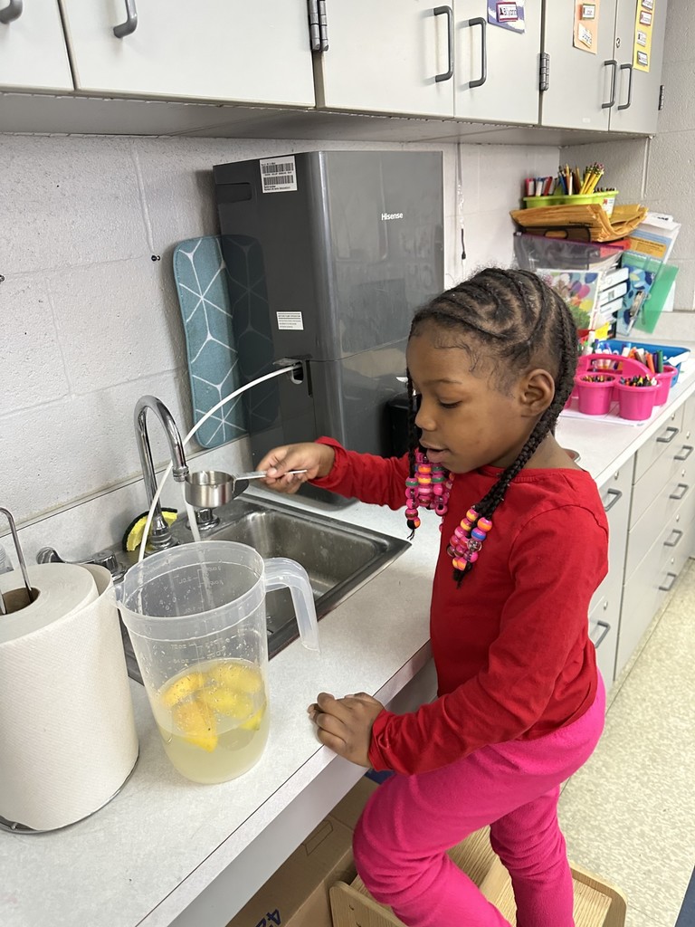 A student is filling a measuring cup with water at a sink with a clear pitcher in a school classroom. Paper towels can be seen nearby.