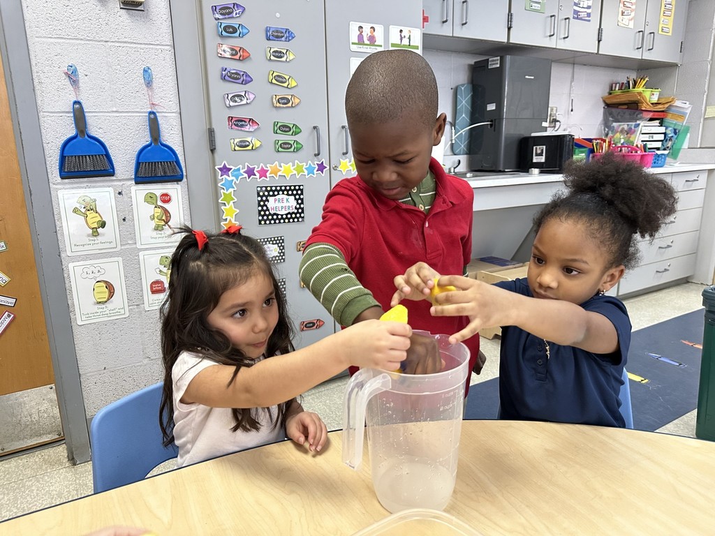 A group of three students are gathered around a table in a school classroom, squeezing lemons into a clear pitcher. Educational posters and storage cabinets can be seen in the background.