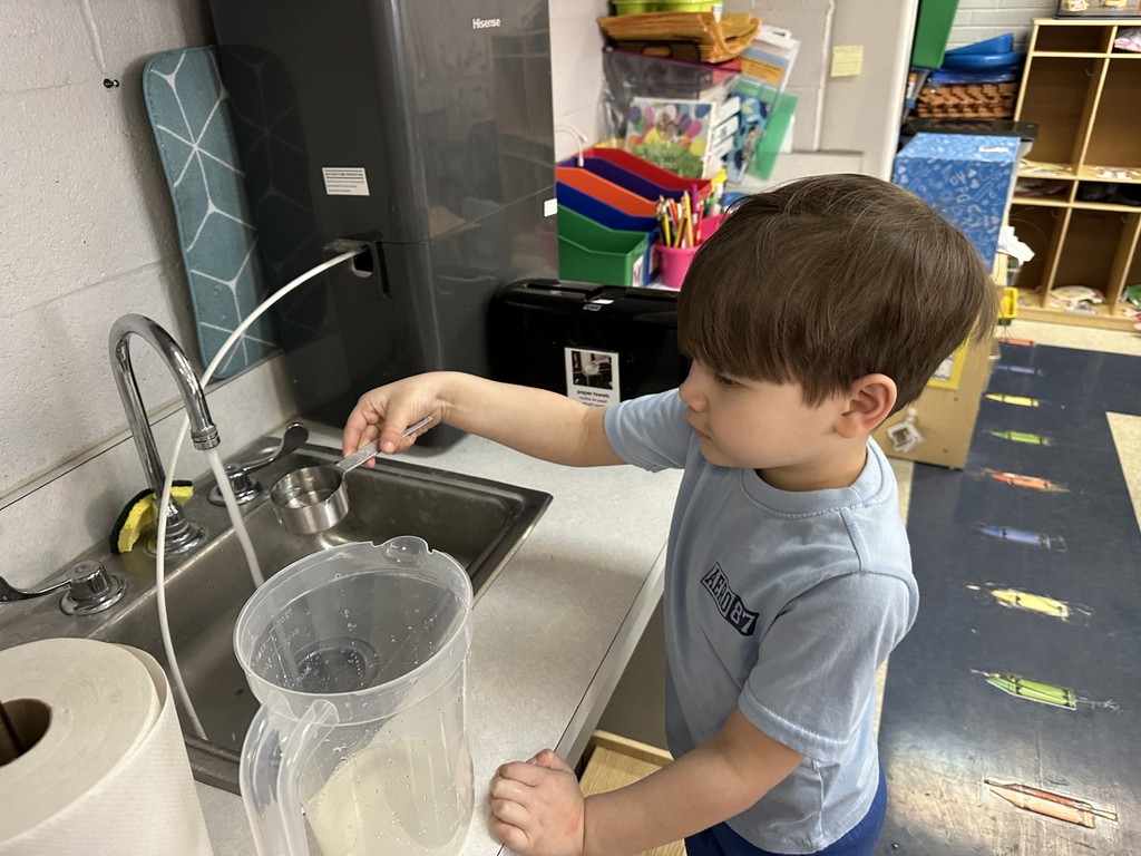 A student is filling a measuring cup with water at a sink with a clear pitcher in a school classroom. Paper towels can be seen nearby.