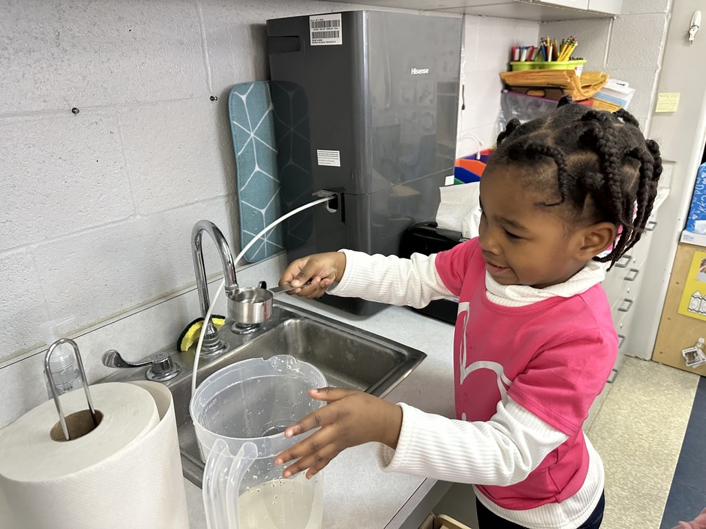 A student is holding a clear pitcher with water in it at a sink. A paper towel roll is nearby. 
