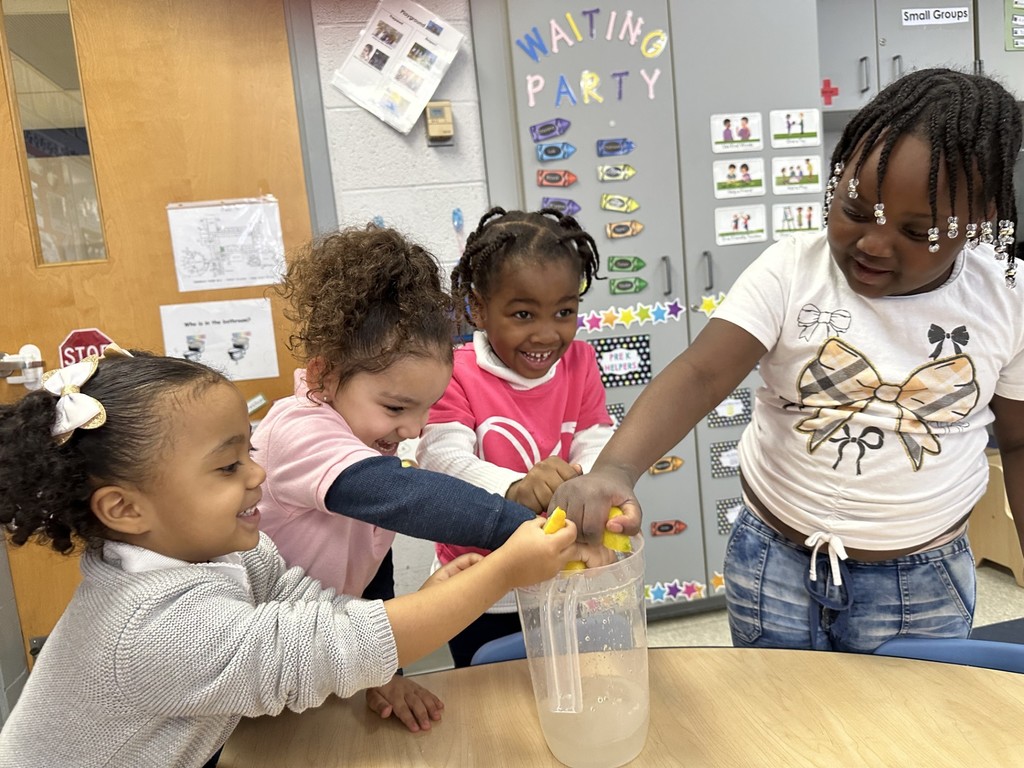 A group of four students are gathered around a table in a school classroom, squeezing lemons into a clear pitcher. Educational posters and storage cabinets can be seen in the background.