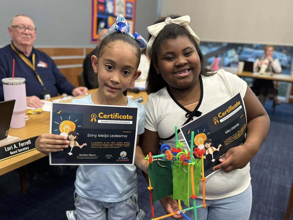 Two students are standing in a conference room holding certificates and a colorful craft project in their hands. Other people can be seen observing in the background.