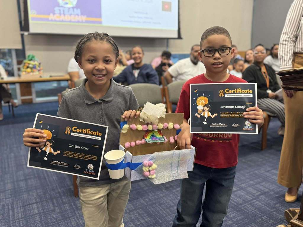 Two students are standing in a conference room holding certificates and a colorful craft project in their hands. A projector screen and other people can be seen in the background.