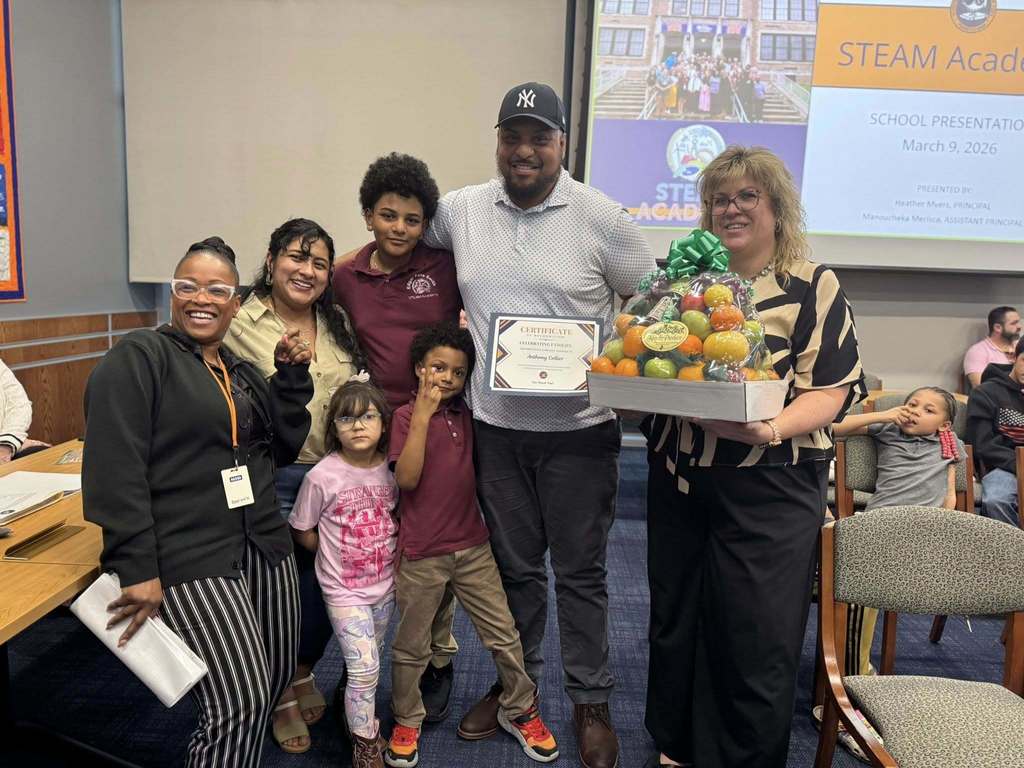 Smith STEAM Academy administrators and a family of five are standing in a conference room. The man is holding a certificate in his hand and a Smith STEAM Academy administrator is holding a fruit basket in her hand. A projector screen and other people can be seen in the background.