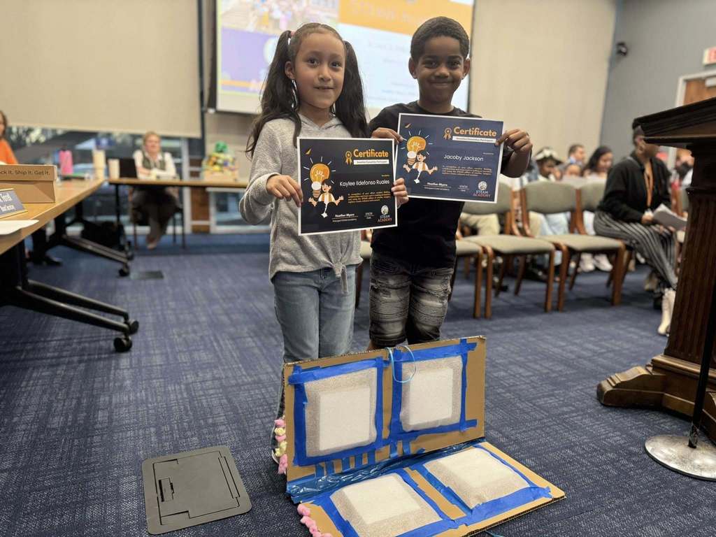 Two students are standing in a conference room holding certificates in their hands. A craft project can be seen on the floor in front of them. A projector screen and other people can be seen in the background.