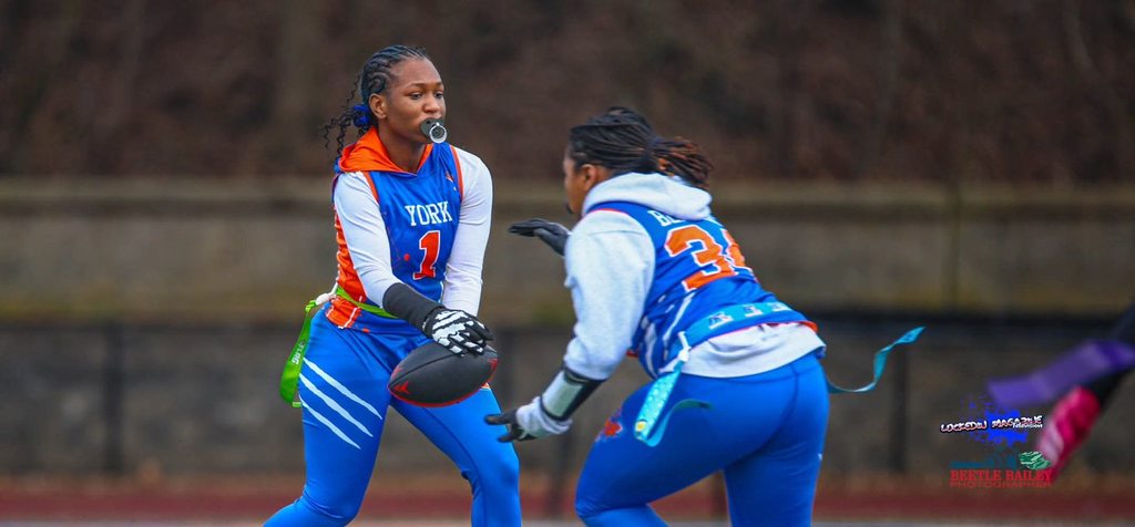 Two young women are playing flag football outdoors. One of the young women is handing off the football to the other young woman.