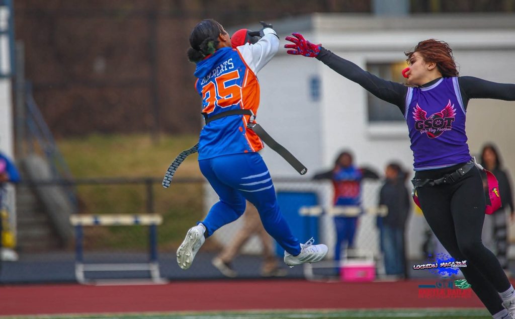 Two young women are playing flag football outdoor in a field. One of the young woman is leaping in the air to intercept a pass, while the other young woman is reaching to block the pass. Other people can be seen observing nearby in the background.