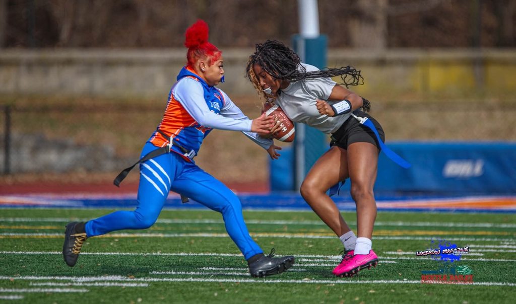 Two young women are playing flag football outdoors on a field. One of the young woman is running with the ball, while the other young woman is attempting a tackle.