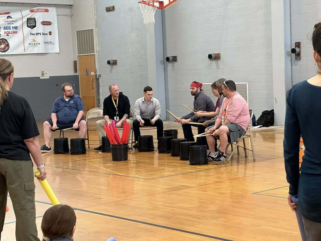 A group of district staff members are sitting on chairs in a school gym playing makeshift drums using buckets and sticks. Other district staff members can be seen observing them.