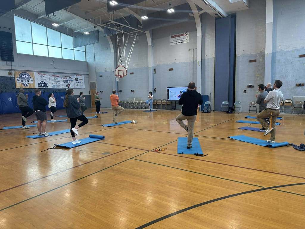A group of district staff members are practicing yoga poses in a school gym. They are spread out on blue mats and an instructor can be seen standing at the front near a large screen.