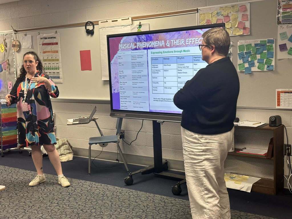 Two district staff members are standing at the front of a school classroom in front of a large screen. The slide, titled "Musical Phenomena & Their Effects," lists various emotions and music elements.