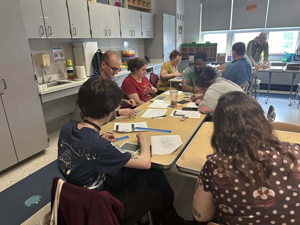 A group of district staff members are engaged in a collaborative activity while sitting around a table in a school classroom. Papers, rulers, and writing tools are scattered.