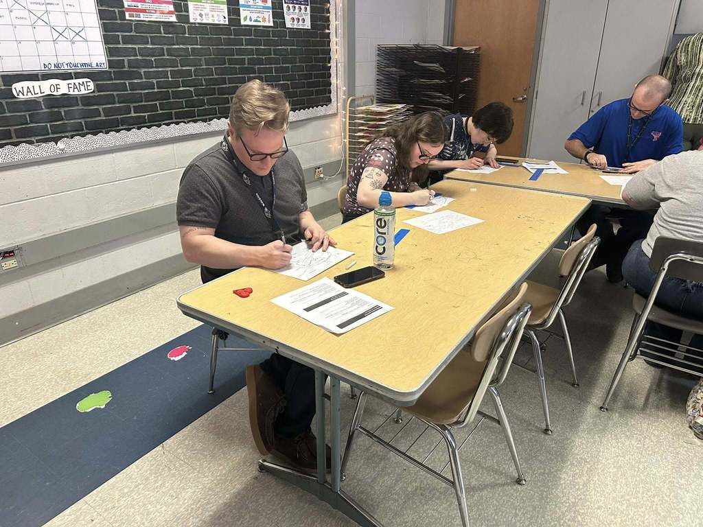A group of district staff members are sitting at a table in a school classroom, focused on drawing. Papers and a water bottle are on the table, with a bulletin board in the background.