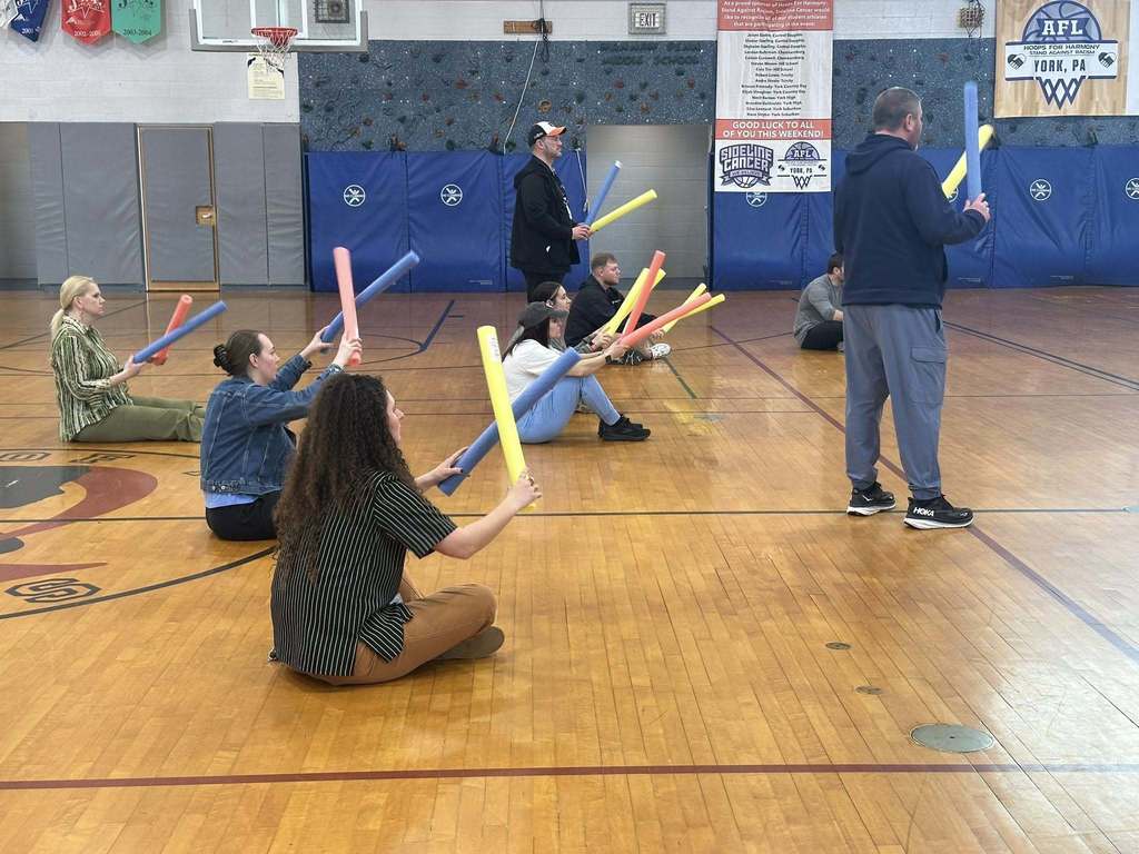 A group of district staff members are sitting and standing on a school gym floor, each of them holding a colorful foam noodle. 