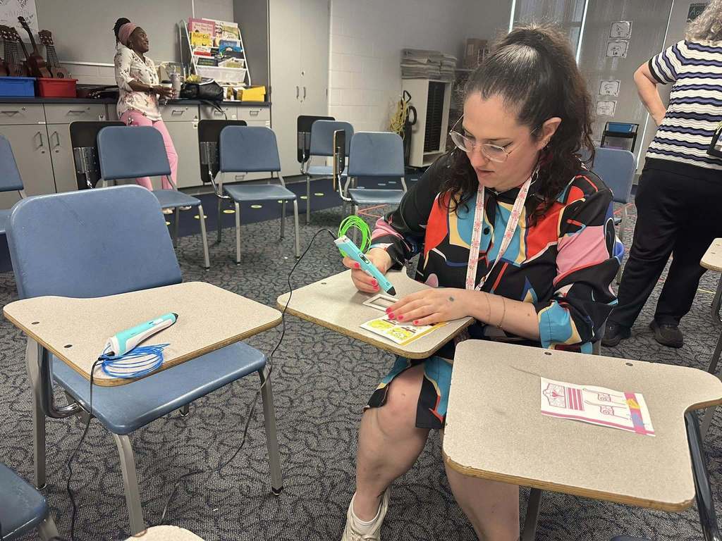 A district staff member is using a 3D pen to trace a drawing while sitting at a desk in a school classroom. Other district staff members can be seen nearby in the background.