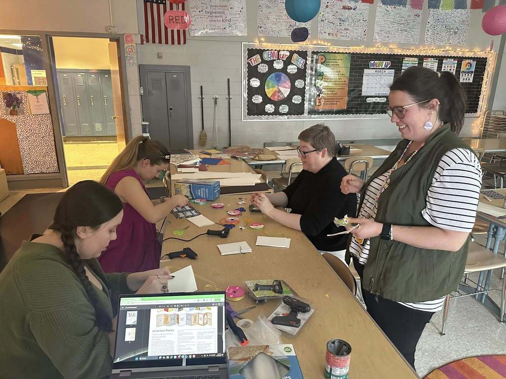 A group of district staff members are sitting around a table in a school classroom engaging in a craft activity using paper and glue guns. A laptop displaying a poetry website can be seen.