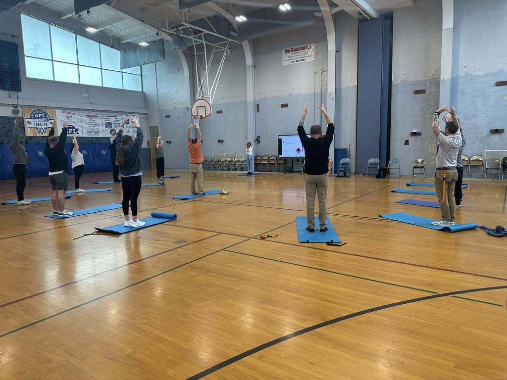 A group of district staff members are practicing yoga poses in a school gym. They are spread out on blue mats and their arms are raised in the air. An instructor can be seen standing at the front near a large screen.