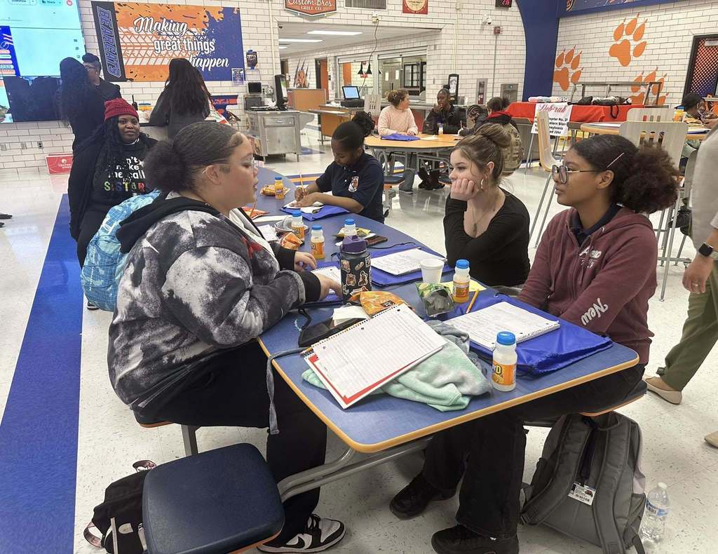 A group of students are sitting at a table in a school cafeteria, engaging in a conversation. Other people can be seen nearby in the background.