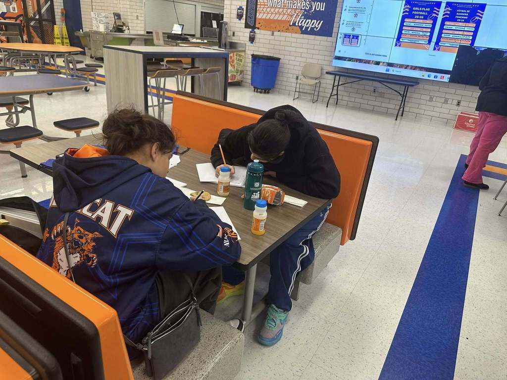 Two students are sitting at a table in a school cafeteria writing on notebooks.