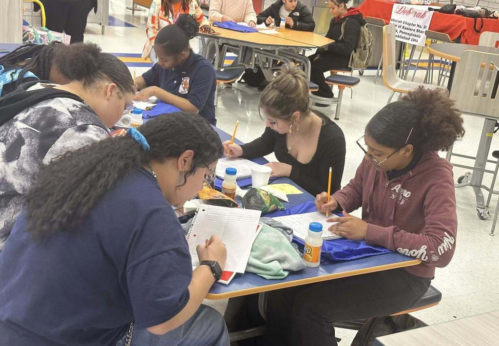 A group of students are sitting at a table in a school cafeteria writing on notebooks. Other people can be seen nearby.