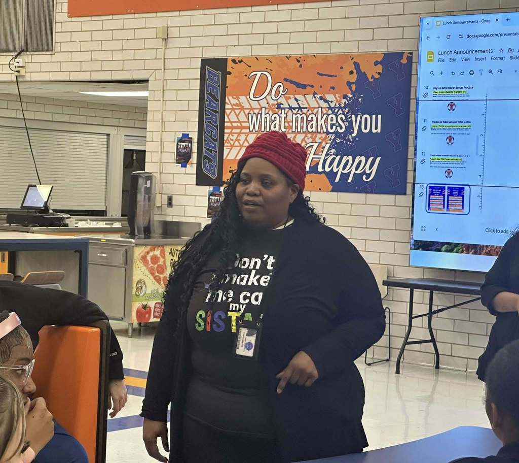School Board Director, Tynisha Wilkes, is standing in a school cafeteria speaking to a group of students. A large monitor is displaying lunch announcements in the background.