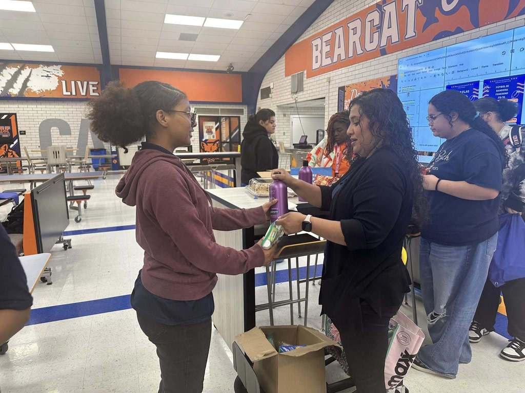 A district staff member is handing some hygiene items to a student in a school cafeteria. Other people can be seen nearby.