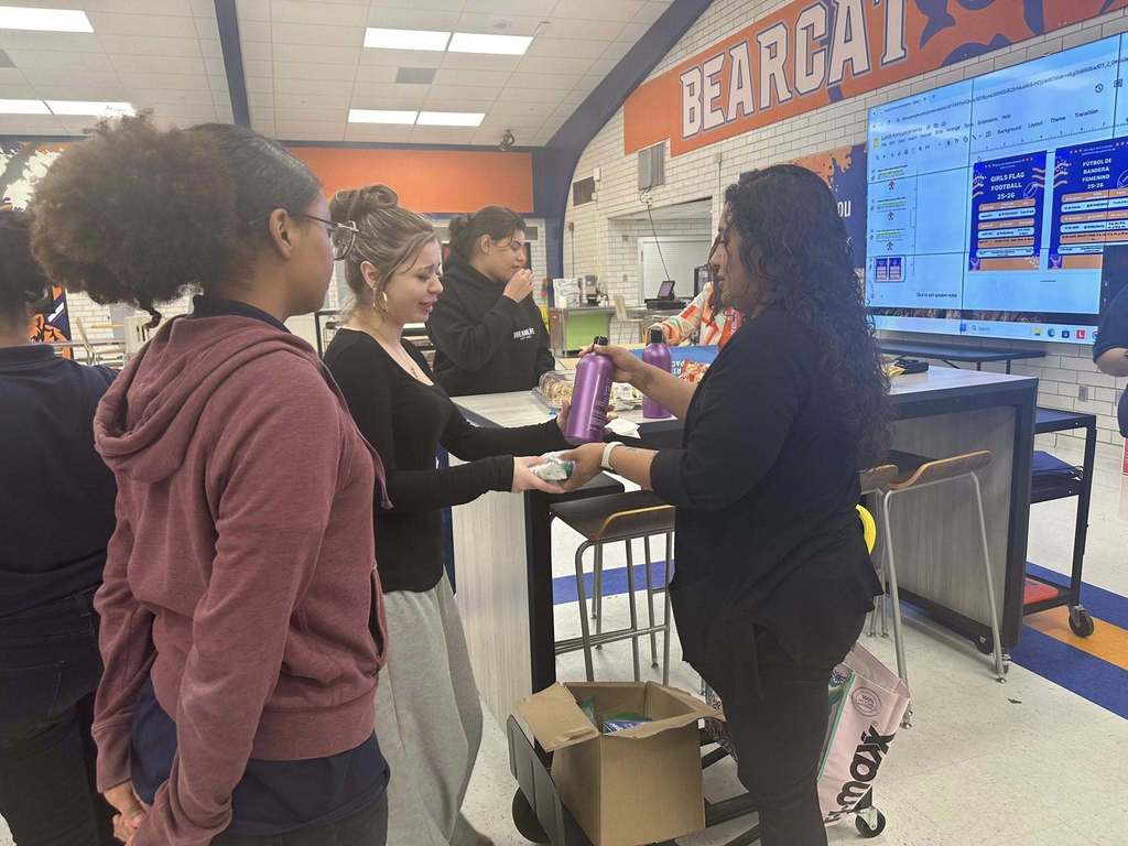 A district staff member is handing some hygiene items to a student in a school cafeteria. Other people can be seen nearby.